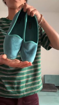 Scarpe slip-on turchesi realizzate in materiale morbido, con una texture liscia e forma arrotondata, tenute in mano contro una camicia a righe verdi.