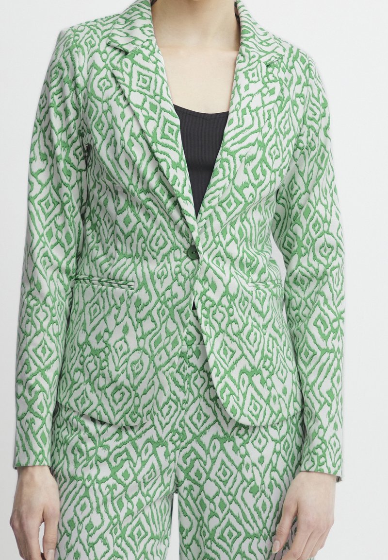 Costume sur mesure avec un motif géométrique vert et blanc, porté sur un haut noir, montrant le torse et les bras sur un fond uni.