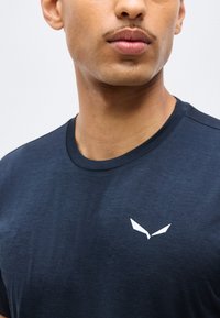 Κοντινό πλάνο ενός ατόμου που φοράει μπλε σκούρο μπλουζάκι με στρογγυλή λαιμόκοψη και μικρό άσπρο γωνιώδες λογότυπο στο αριστερό στήθος.