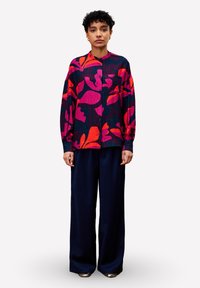 Femme avec les cheveux courts et bouclés portant une chemise à manches longues à motifs floraux colorés et un pantalon large bleu marine, se tenant devant un fond blanc.