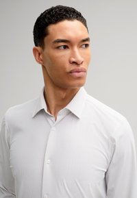 Camicia bianca con bottone, caratterizzata da un motivo a pois sottile, colletto classico e texture del tessuto liscia. Il modello mostra un'espressione neutra.