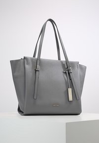Sac fourre-tout en cuir gris avec deux longues poignées, une forme structurée, une surface texturée, un logo en métal en accent, et une étiquette amovible.