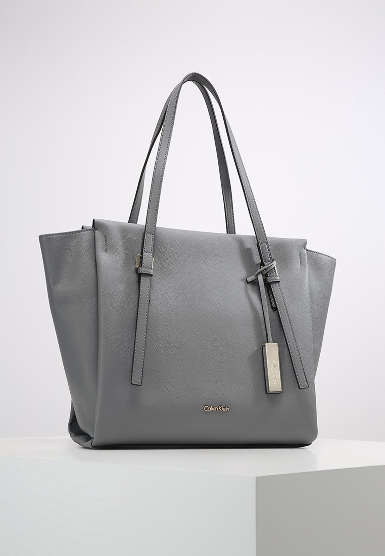 Sac fourre-tout en cuir gris avec deux longues poignées, une forme structurée, une surface texturée, un logo en métal en accent, et une étiquette amovible.