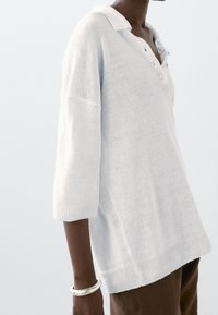 Weißer gestrickter Pullover mit Kragen, Dreiviertelärmeln und geripptem Saum. Mit zwei Knöpfen am Kragen. Weiche Textur.