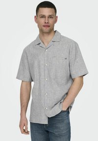 Korte mouwen, gekraagde shirt in het grijs met witte strepen. Voorzien van een linkse borstzak en een knoopsluiting aan de voorkant. Gedragen met een blauwe spijkerbroek.