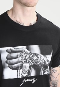 Black cotton t-shirt featuring a large black-and-white graphic of a hand adorned with jewelry and intricate tattoos. Text reads "pray."