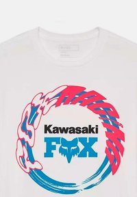 Camiseta blanca de algodón con un gráfico circular en azul y rosa. El texto central dice "Kawasaki" sobre "FOX" con un logo de zorro estilizado.