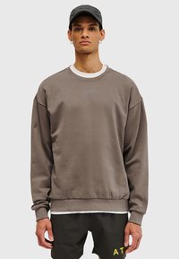 Brun crewneck-tröja med vit innerkrage, med ordet "HALO" i ljusblått på bröstet. Mjukt tyg och avslappnad passform.