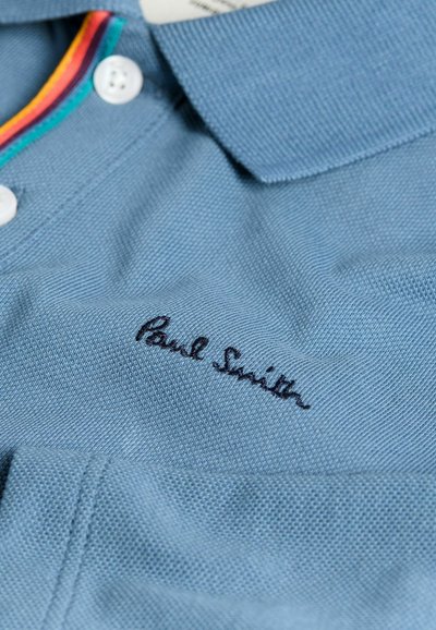 Šviesiai mėlynas polo marškinėliai, pagaminti iš tekstūruoto audinio, su apykakle, dekoruota raudonais, geltonais ir žaliais dryžiais, ir juodu siuvinėtu "Paul Smith" logotipu.