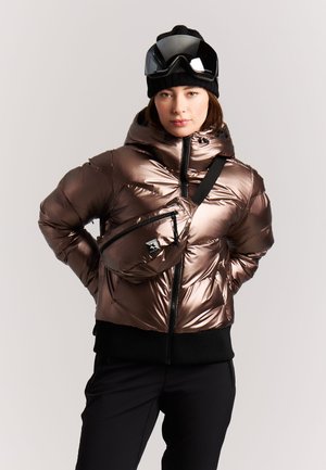 Vrouw draagt glanzende bronzen donzen jas, zwarte broek, zwarte muts met skibril en een bijpassende bronzen crossbody tas tegen een effen achtergrond.