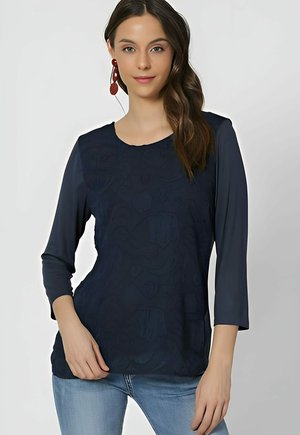 Camisa azul marino de mangas tres cuartos, con patrones texturizados en la parte frontal y un escote redondo, combinada con jeans de color azul claro.