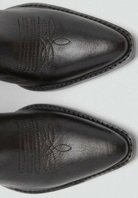 Botas de cuero negro con punta afilada, que presentan costuras y patrones florales grabados en la parte superior. Textura duradera y suave, con un borde detallado.
