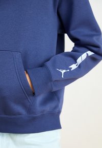 Sweatshirt capuz azul-marinho com um bolso canguru, punhos canelados e um logótipo gráfico branco na manga. O tecido parece suave e macio.
