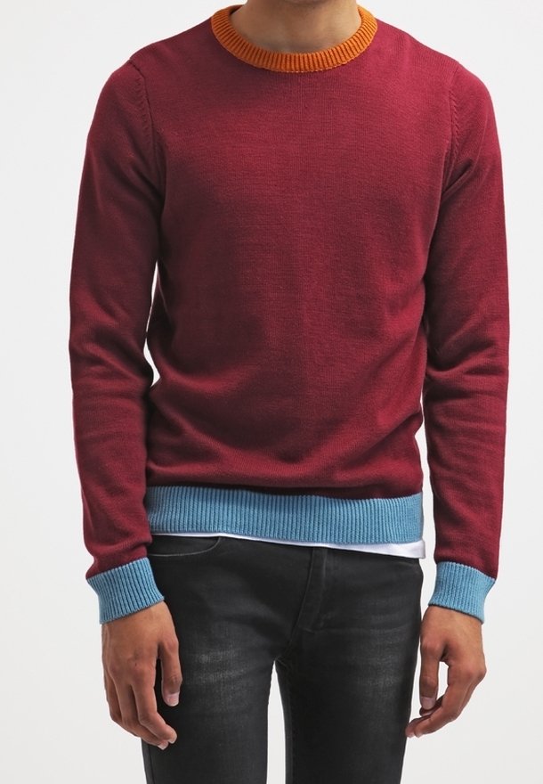 Homme portant un pull bordeaux à manches longues avec un col orange, des poignets et un ourlet bleus, associé à un jean noir, debout les bras détendus.