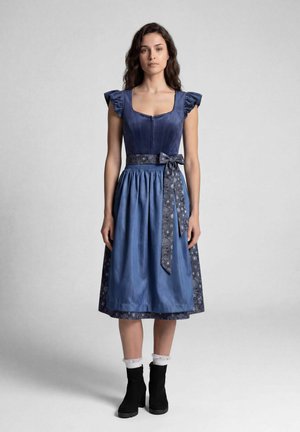 Frau, die ein blaues traditionelles Kleid mit einem floralen Taillengürtel und schwarzen Knöchelstiefeln mit weißen Rüschenstrümpfen trägt, steht vor einem neutralen Hintergrund.