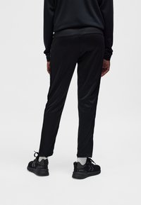 Eine Person trägt schwarze Sporthosen, schwarze Turnschuhe mit weißen Socken und steht mit dem Rücken zur Kamera vor einem einfarbigen hellen Hintergrund.