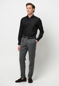 Camicetta nera a maniche lunghe, pantaloni grigi con texture, cintura in pelle marrone e scarpe scamosciate marroni scure, in piedi davanti a uno sfondo bianco semplice.