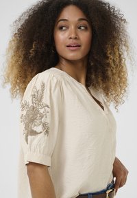 Blouse beige en tissu texturé, avec des broderies florales sur la manche gauche, des manches courtes évasées et un décolleté en V.