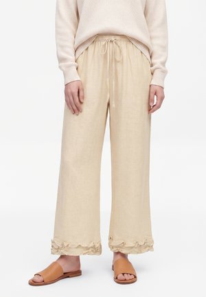 Beige wijde broek met ruchtrand aan de zoom en een koordje in de taille, gecombineerd met bruine slides en een licht gestructureerde trui.