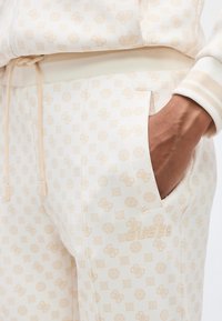 Χέρι ατόμου στην τσέπη κρεμ χοντρού φούτερ με σχέδια και ζωνάκι κορδονιών στη μέση, συνοδευόμενο από παρόμοιο φούτερ με σχέδια.