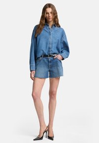 Camicia di jeans con bottone frontale e vestibilità oversize, abbinata a pantaloni corti di jeans azzurri con bordi sfrangiati e scarpe nere con tacco alto.