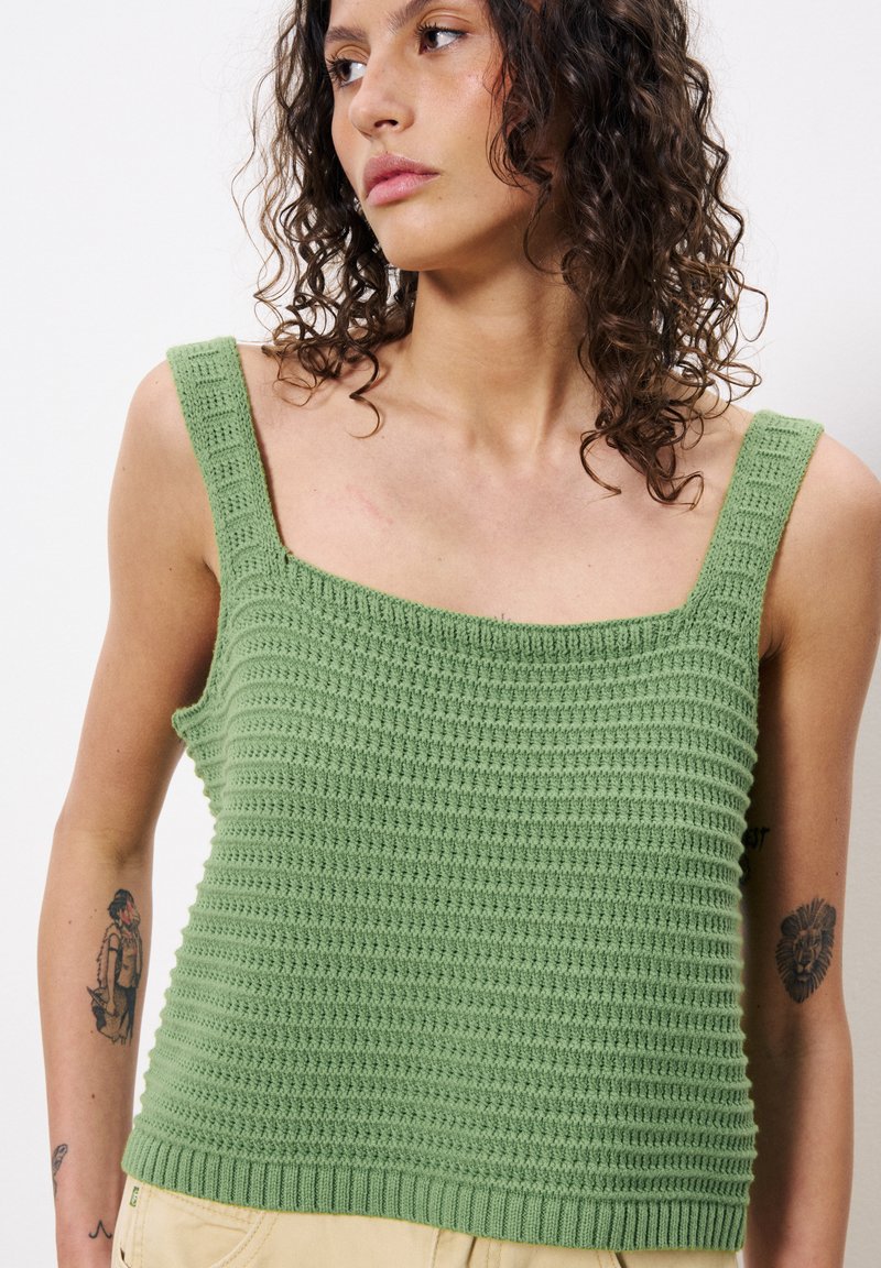 Femme aux cheveux bouclés portant un débardeur en maille vert et un pantalon beige, affichant des tatouages sur les deux bras sur fond blanc.