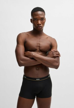 Schwarze Boxershorts mit einem goldenen Akzentbund, der das "BOSS"-Logo trägt. Glattes Material, enganliegendes Design und Oberschenkel-lange Länge. Haut ist sichtbar darüber.