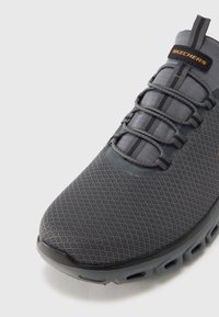 Zapato deportivo gris con parte superior de malla, cordones elásticos y un logo prominente. La suela cuenta con secciones acolchadas y orificios de ventilación.