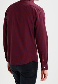 Camicia a maniche lunghe di color rosso scuro con motivo a quadri blu navy, colletto classico, polsini con bottone e orlo dritto. Materiale in misto cotone.