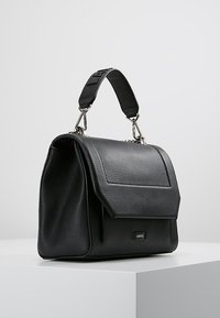 Sac à main en cuir noir avec une surface texturée, un rabat angulaire, des accessoires en métal et une bandoulière amovible. Marque affichée à l'avant.