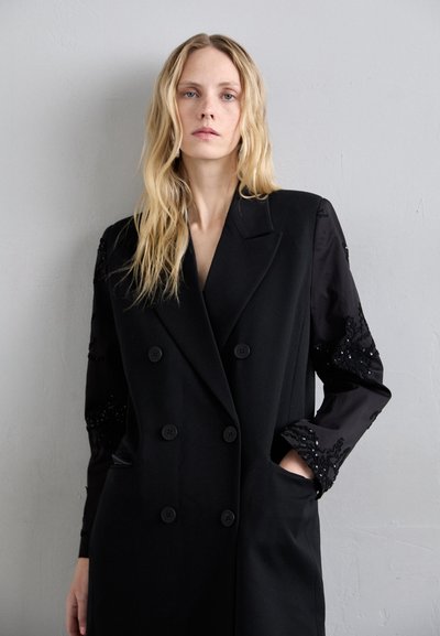 Melnā divkrūšu mētelis ar teksturētām piedurknēm, kas rotāts ar melnām sequīnām, ar pielāgotu dizainu un priekšējām kabatām. Gluda auduma tekstūra.