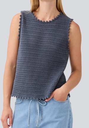 Ärmelloses dunkelgraues gestricktes Top mit welligen Rändern, getragen von einer Person mit hellblauer Jeans, eine Hand in der Tasche.