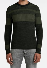 Zielony sweter dzianinowy z różnorodnymi fakturami i poziomymi paskami. Prążkowany dekolt w serek i mankiety. Noszony z szarymi dżinsami.