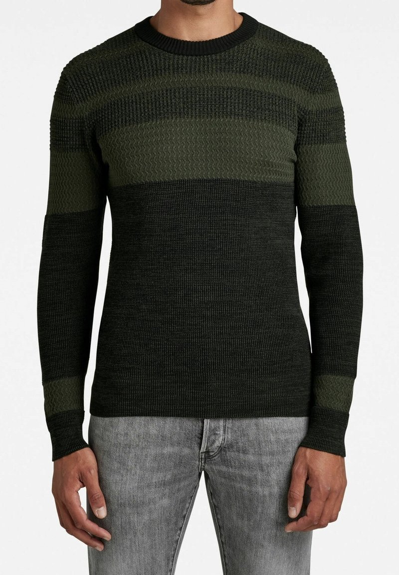 Zielony sweter dzianinowy z różnorodnymi fakturami i poziomymi paskami. Prążkowany dekolt w serek i mankiety. Noszony z szarymi dżinsami.