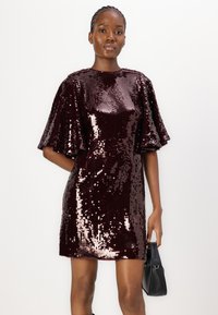 Robe en sequins bordeaux avec des manches courtes et évasées. Surface texturée avec une finition scintillante. Associée à un petit sac à main noir.