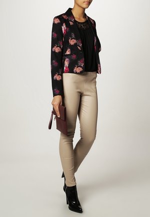Vrouw draagt beige leren broek, zwarte blazer met bloemenmotief, zwart topje, zwarte enkellaarzen en houdt een bordeauxrode clutch vast.