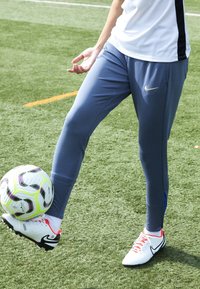 Blauwe sportbroek met een aansluitend ontwerp en zijaccentdetails, gecombineerd met witte sportschoenen met rode veters, op kunstgras.