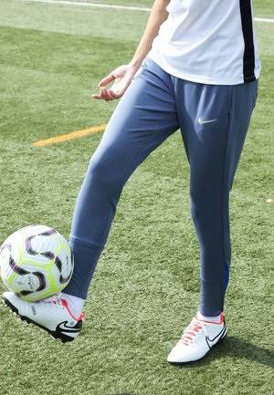 Niebieskie sportowe spodnie o dopasowanym kroju z bocznymi detalami, w zestawieniu z białymi butami sportowymi z czerwonymi sznurówkami, na sztucznej murawie.