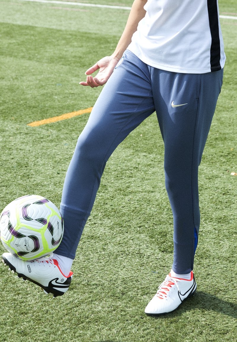 Blauwe sportbroek met een aansluitend ontwerp en zijaccentdetails, gecombineerd met witte sportschoenen met rode veters, op kunstgras.