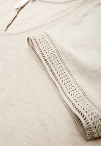 Beige v-neck garment with textured fabric and decorative braided trim along the sleeve edge on a flat surface.
