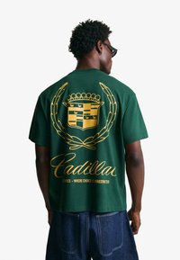 Green cotton T-shirt featuring a large gold emblem and script text on the back. Design includes a crown, laurel leaves, and CADILLAC branding.