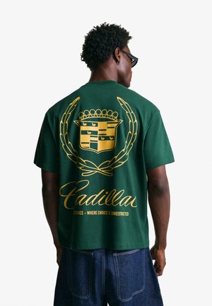Green cotton T-shirt featuring a large gold emblem and script text on the back. Design includes a crown, laurel leaves, and CADILLAC branding.