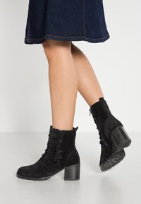 Bottines en daim noir avec un talon empilé, détail de frange et fermeture éclair sur le côté ; texture douce et design structuré.