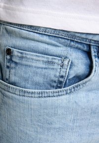 Svetle modré džíny z denimu s přední kapsou, s prošívanými švy a malým černým knoflíkem. Látka má vybledlou texturu.