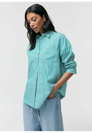 Kobieta ubrana w zielono-białą koszulę z pionowymi paskami, z podwiniętymi rękawami, oraz jasnoniebieskie dżinsy, z ręką w kieszeni, patrząca w dół.