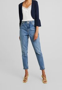 Un cardigan court marine sur un camisole blanc à dentelle, associé à un jean taille haute bleu clair et des talons jaunes à lanières.