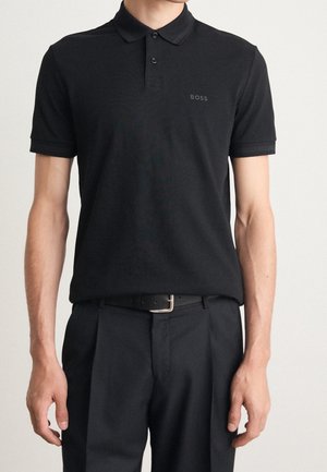 Mann trägt ein schwarzes Polohemd mit kurzen Ärmeln und einem kleinen "BOSS" Logo auf der Brust sowie schwarze Hose mit Gürtel.
