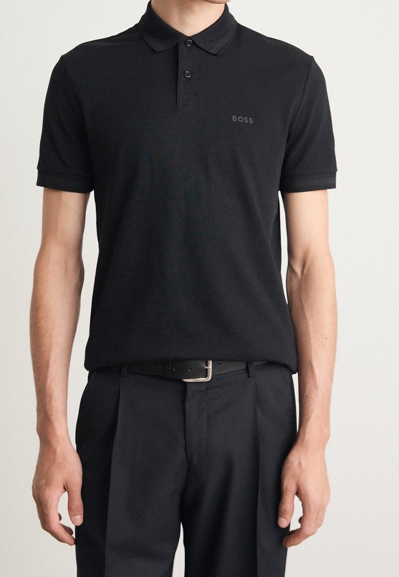Homme portant un polo noir à manches courtes avec un petit logo "BOSS" sur la poitrine et un pantalon noir ceinturé.