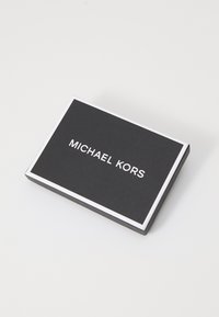 Fekete téglalap alakú doboz texturált felülettel, fehér szélekkel, és a tetején a "MICHAEL KORS" felirat dőlt, fehér betűkkel.