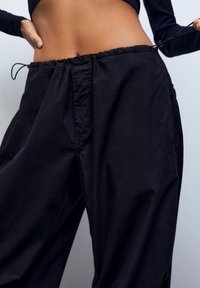 Zwarte, losse broek met een elastische tailleband en een trekkoorddetail. De stof heeft een gladde textuur en een licht gekreukt uiterlijk.
